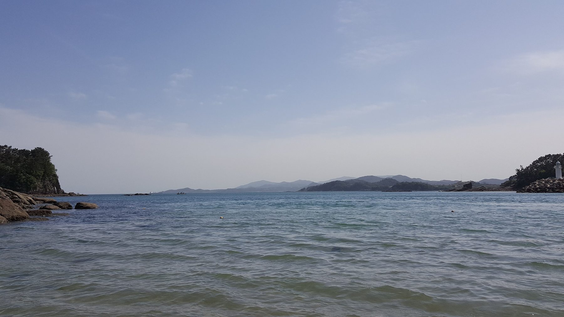 맑은 날의 사천 앞바다. 왼쪽 앞에 소나무가 자란 바위 곶이 있고, 잔물결이 이는 바다 건너 멀리 낮은 산줄기와 섬들이 이어진다. 오른쪽 끝 바위 위에 흰 등대가 서 있다.
