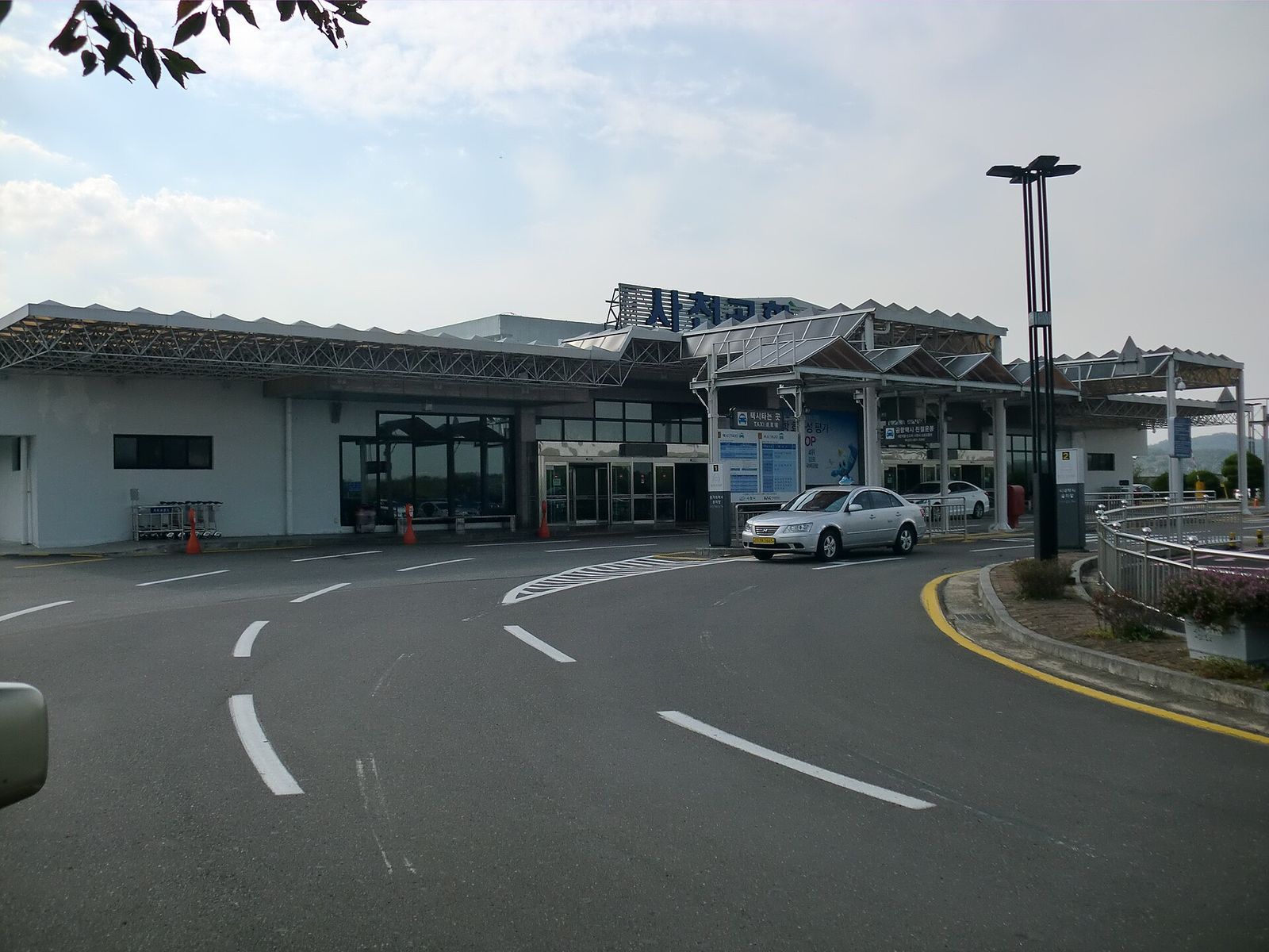 사천공항 여객터미널 바깥을 찍은 사진. 톱날 모양 지붕을 얹은 낮고 흰 건물에 '사천공항' 간판이 붙어 있고, 택시 승강장에 은색 택시 한 대가 서 있다. 앞쪽 차도에는 차선만 그려진 채 비어 있다.