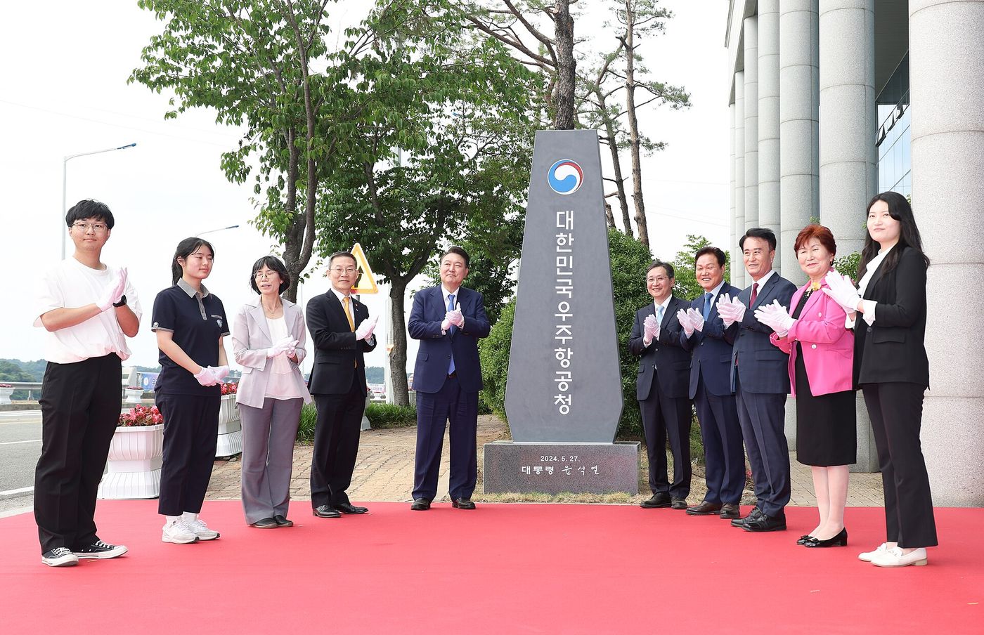 야외 행사장에서 정장과 평상복을 입은 사람들이 흰 장갑을 끼고 박수를 치며 서 있다. 가운데에 정부 상징이 새겨진 검은 돌 표지석이 세워져 있고 '대한민국 우주항공청'이라는 글씨가 세로로 적혀 있다. 발밑에는 붉은 카펫이 깔려 있다.