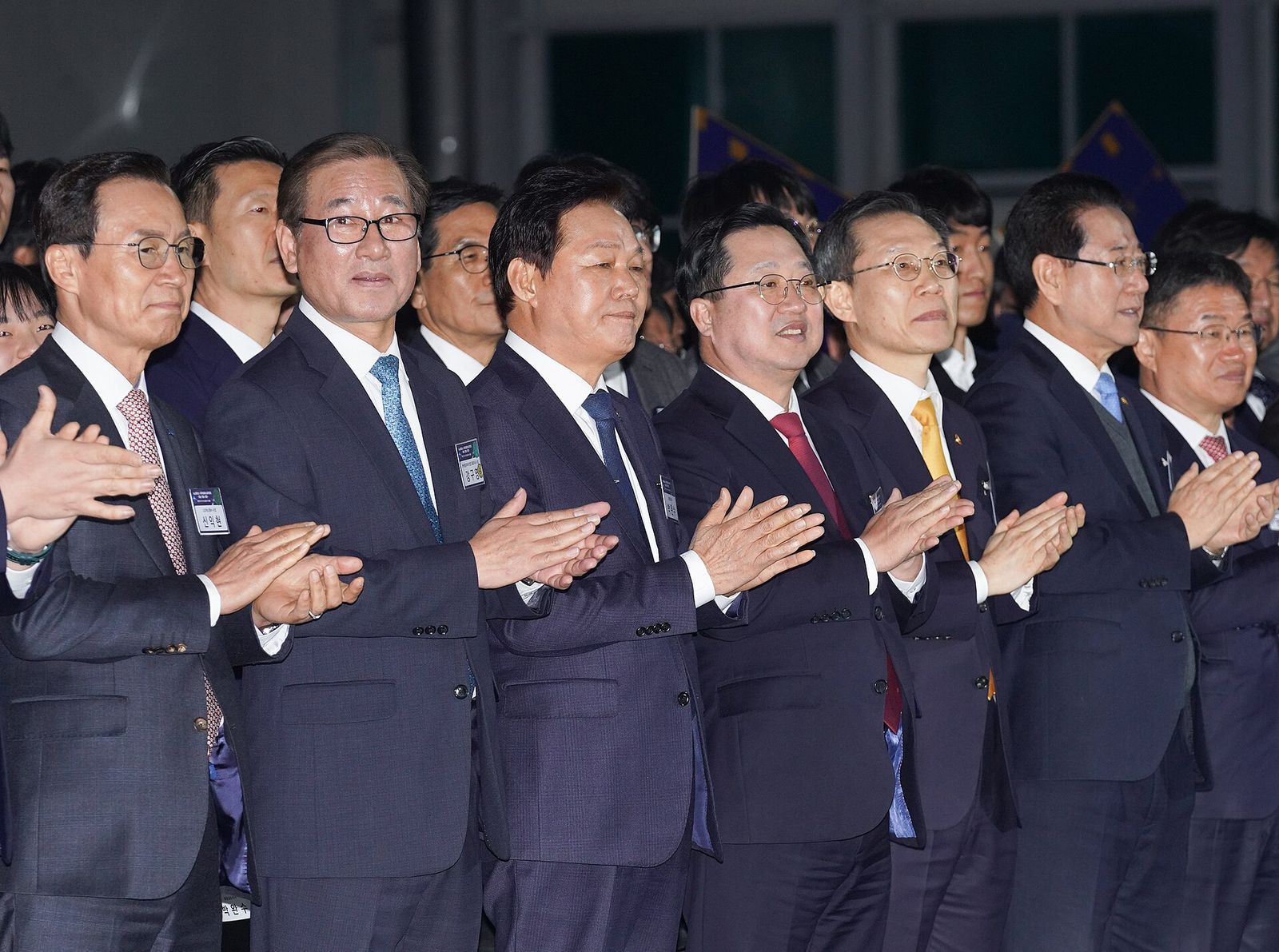 어두운 실내 행사장에서 정장을 입은 사람들이 한 줄로 서서 박수를 치고 있다. 가슴에 이름표를 달고 있고 뒤쪽에 행사용 깃발이 보인다.
