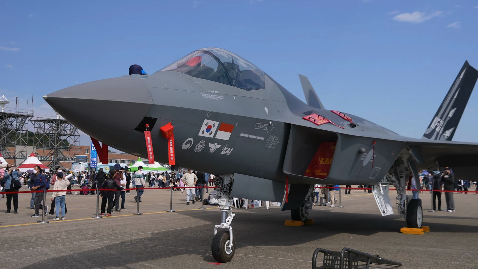 회색 KF-21 전투기가 행사장 바닥에 전시돼 있다. 붉은 줄 뒤로 관람객들이 사진을 찍고 있고, 기체 앞쪽에는 붉은 'REMOVE BEFORE FLIGHT' 표찰이 여러 개 달려 있다. 동체에 태극기와 인도네시아 국기, KAI 표시가 나란히 붙어 있다.