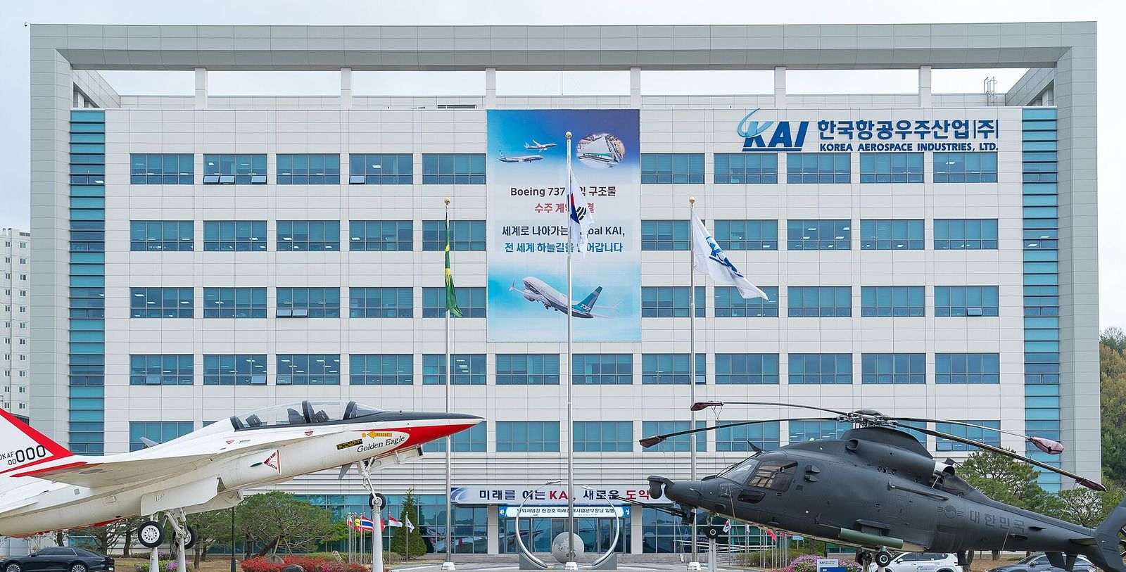 여러 층으로 된 흰 사무동 정면. 벽에 'KAI 한국항공우주산업(주) KOREA AEROSPACE INDUSTRIES, LTD.' 간판이 붙어 있고 가운데에 큰 현수막이 걸려 있다. 건물 앞 광장에는 흰색과 붉은색 T-50 훈련기와 짙은 회색 헬기가 전시대에 올려져 있다.